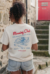 A lady wearing a natural printed graphic t-shirt, with a illustration of a breakfast fry up, set in a town.