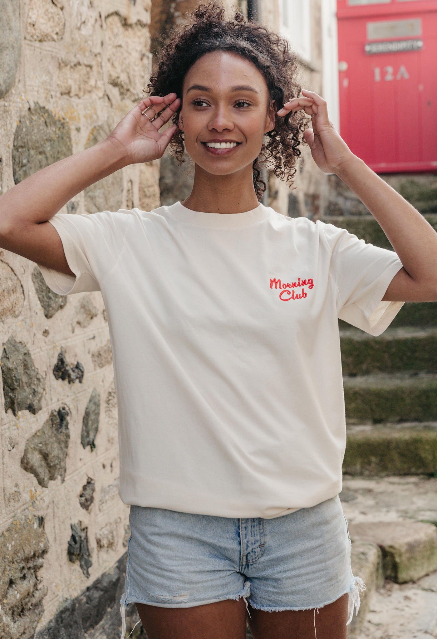 A lady wearing a natural printed graphic t-shirt, with a illustration of a breakfast fry up, set in a town.