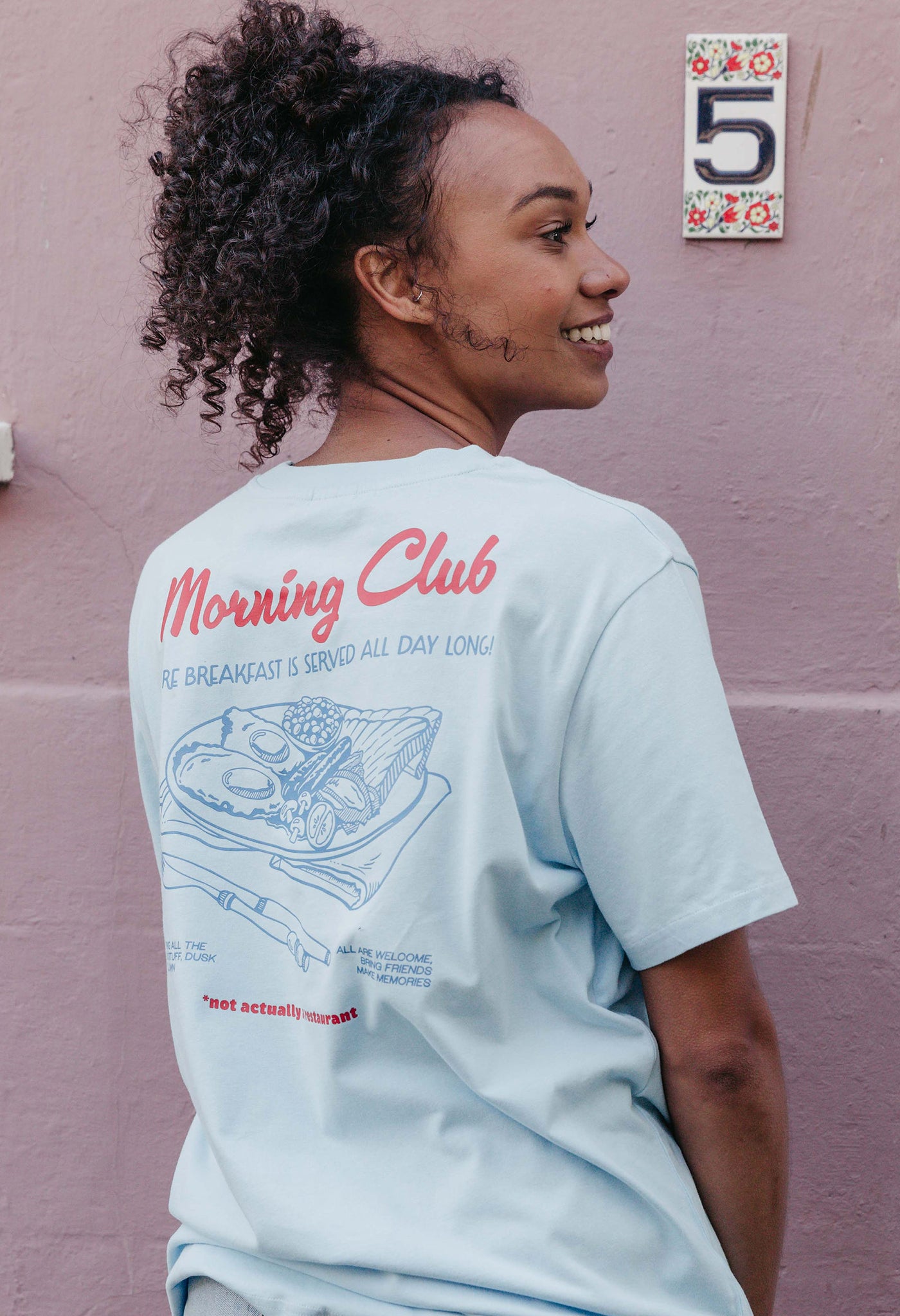 A lady wearing a blue printed graphic t-shirt, with a illustration of a breakfast fry up, set in a town.