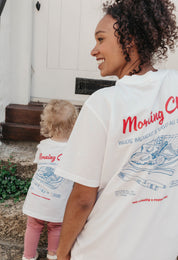 A lady and baby wearing white printed graphic t-shirts, with a illustration of a breakfast fry up, set in a town.