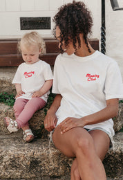 A lady and baby wearing white printed graphic t-shirts, with a illustration of a breakfast fry up, set in a town.