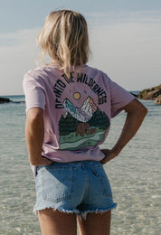 A lady wearing a lilac printed graphic t-shirt, that says Off Into The Wilderness, set by the beach.