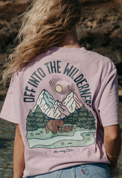 A lady wearing a lilac printed graphic t-shirt, that says Off Into The Wilderness, set by the beach.