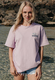 A lady wearing a lilac printed graphic t-shirt, that says Off Into The Wilderness, set by the beach.