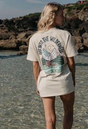 A lady wearing a natural printed graphic t-shirt, that says Off Into The Wilderness, set by the beach.