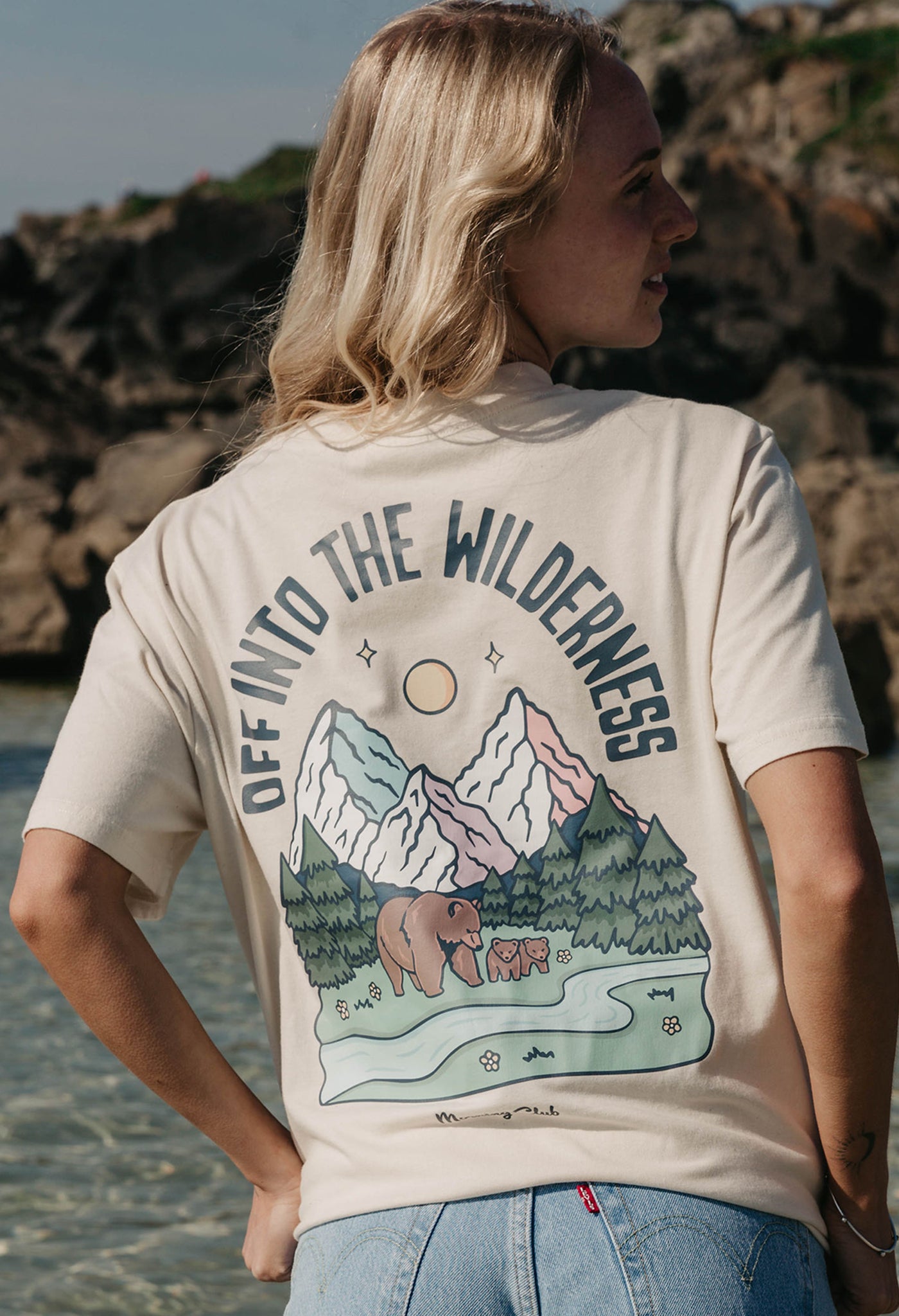 A lady wearing a natural printed graphic t-shirt, that says Off Into The Wilderness, set by the beach.