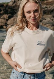 A lady wearing a natural printed graphic t-shirt, that says Off Into The Wilderness, set by the beach.