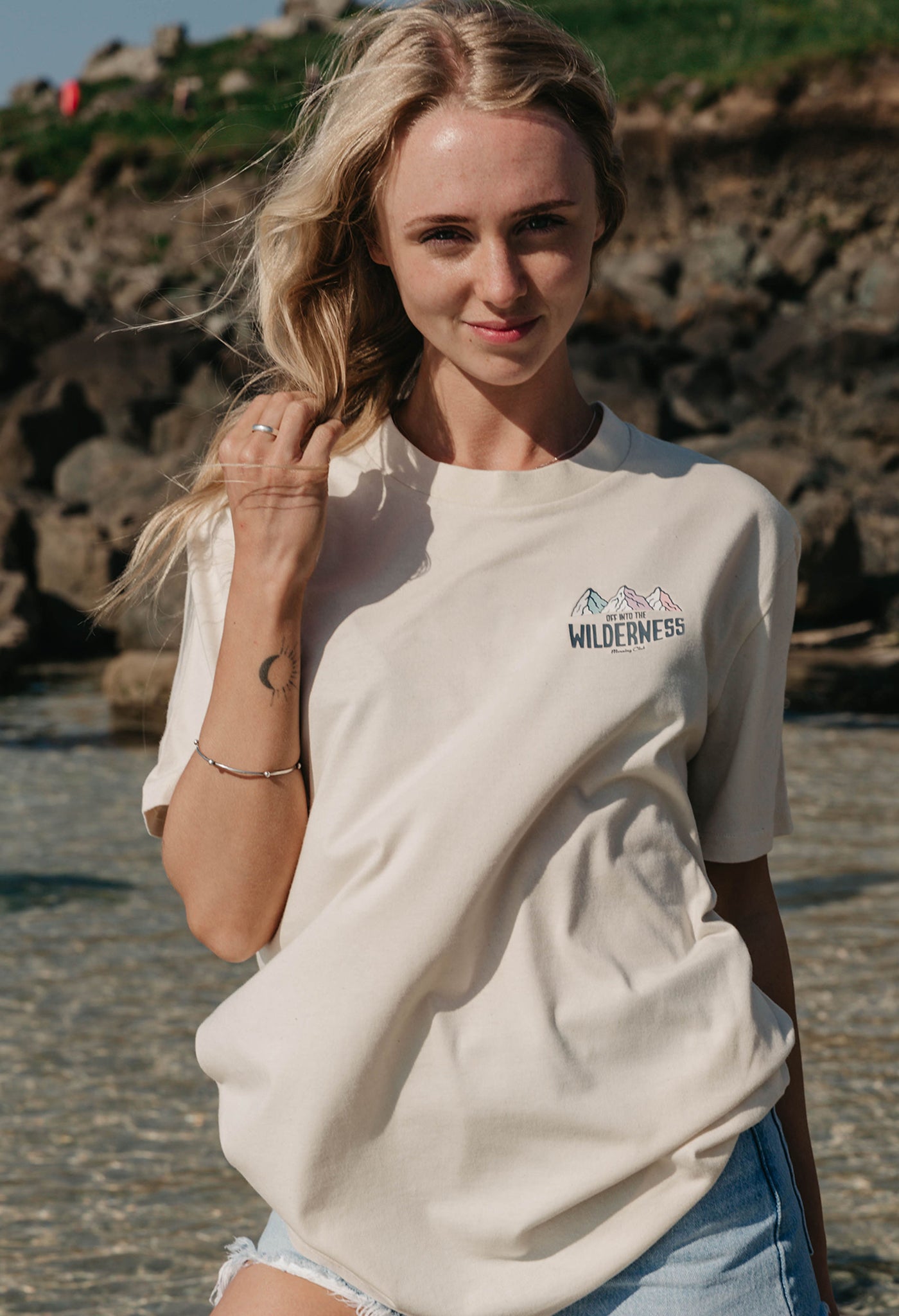 A lady wearing a natural printed graphic t-shirt, that says Off Into The Wilderness, set by the beach.