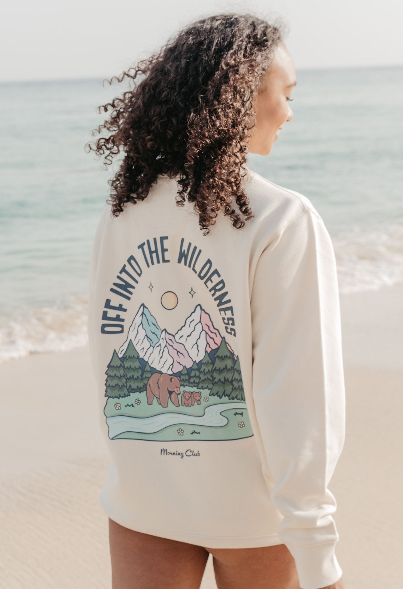 Lady wearing a printed sweatshirt, that says into the wilderness, and its set on the beach.