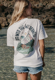 A lady wearing a white printed graphic t-shirt, that says Off Into The Wilderness, set by the beach.