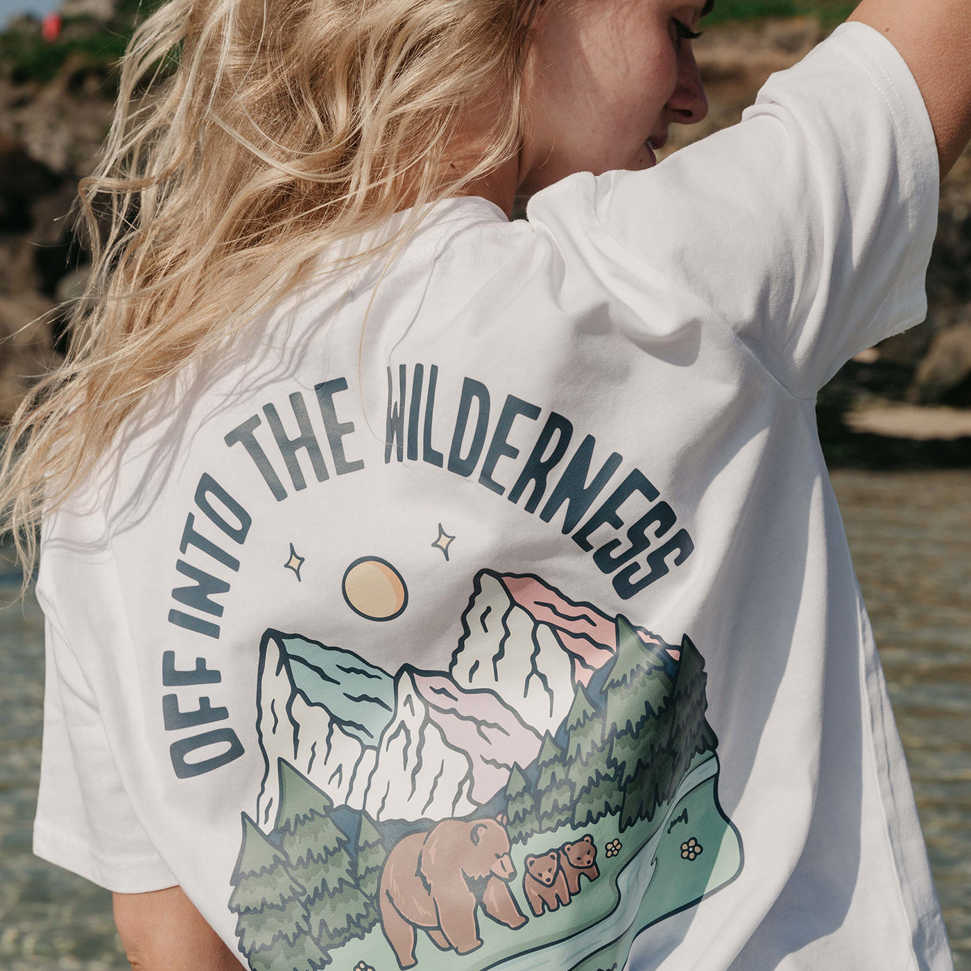 A lady wearing a white printed graphic t-shirt, that says Off Into The Wilderness, set by the beach.