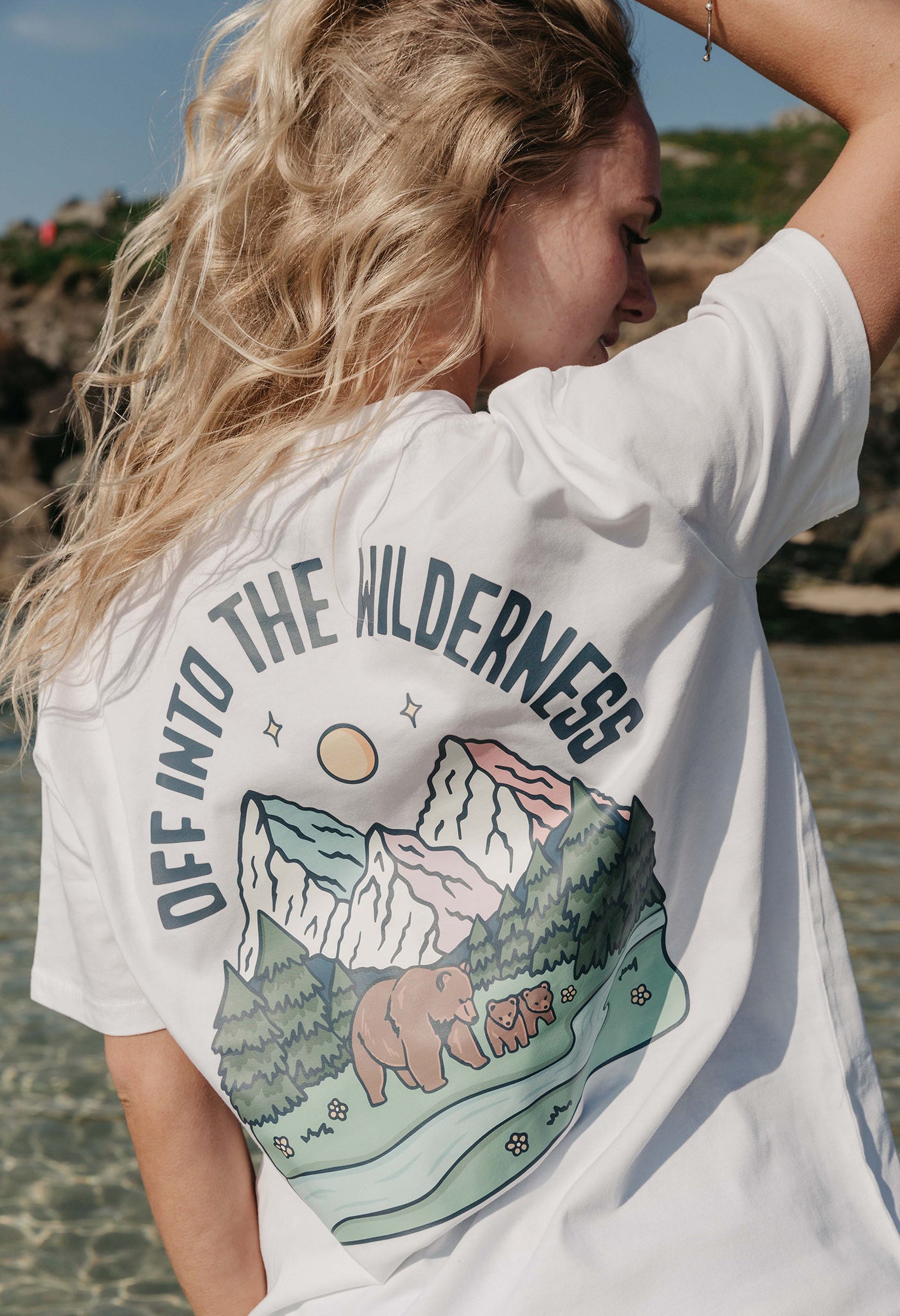 A lady wearing a white printed graphic t-shirt, that says Off Into The Wilderness, set by the beach.