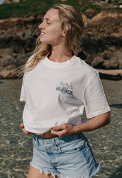 A lady wearing a white printed graphic t-shirt, that says Off Into The Wilderness, set by the beach.