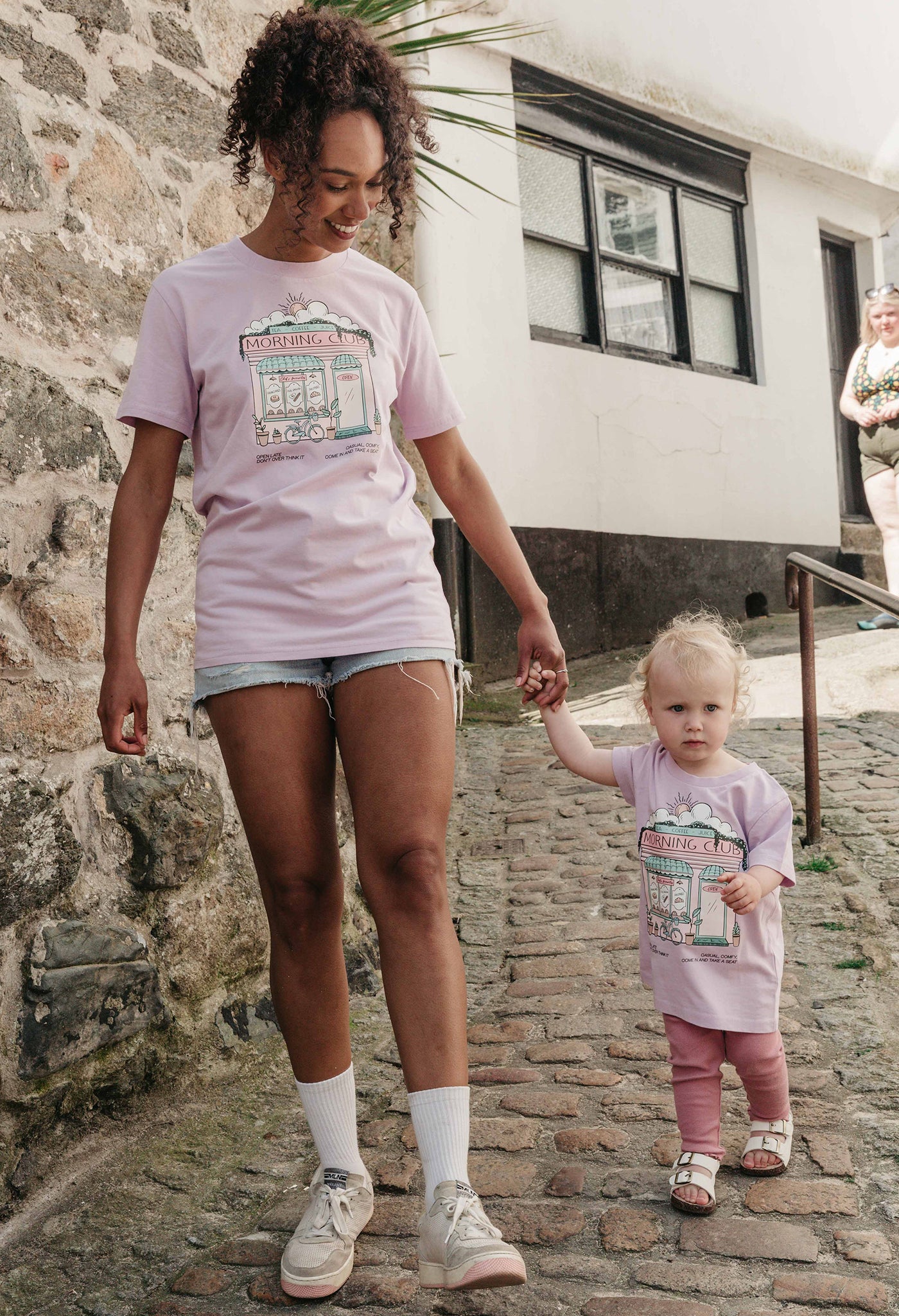 A lady and baby wearing a lilac printed graphic t-shirt, Morning Club Cafe, set in a town.