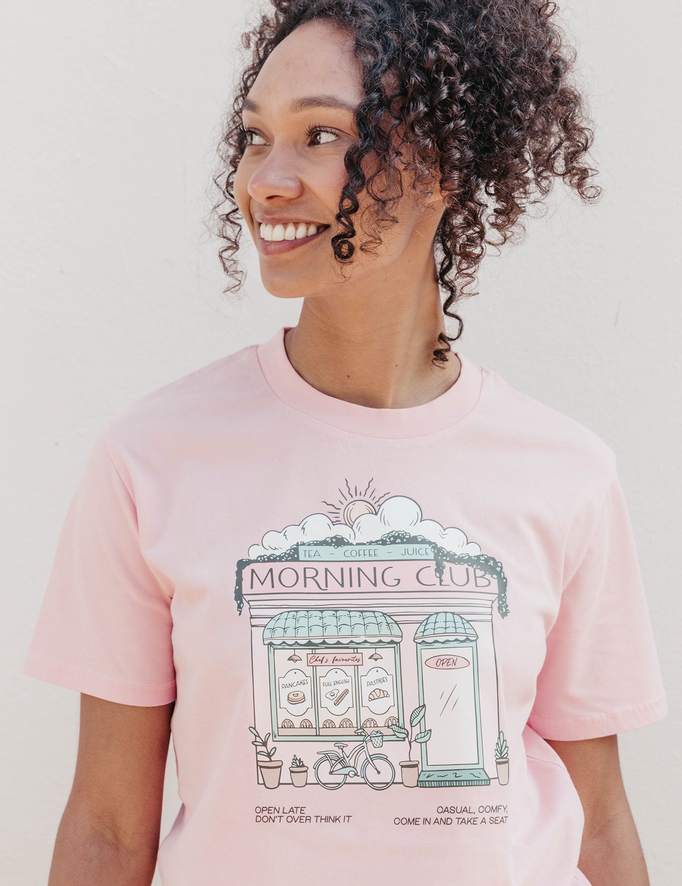 A lady wearing a pink printed graphic t-shirt, Morning Club Cafe, set in a town.