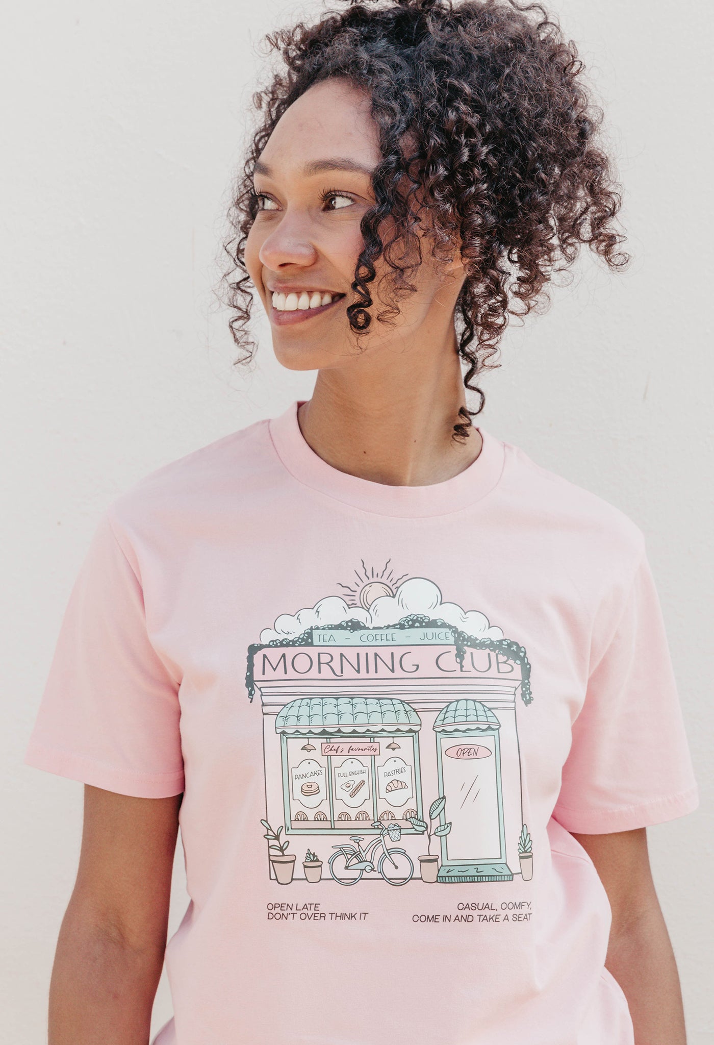 A lady wearing a pink printed graphic t-shirt, Morning Club Cafe, set in a town.