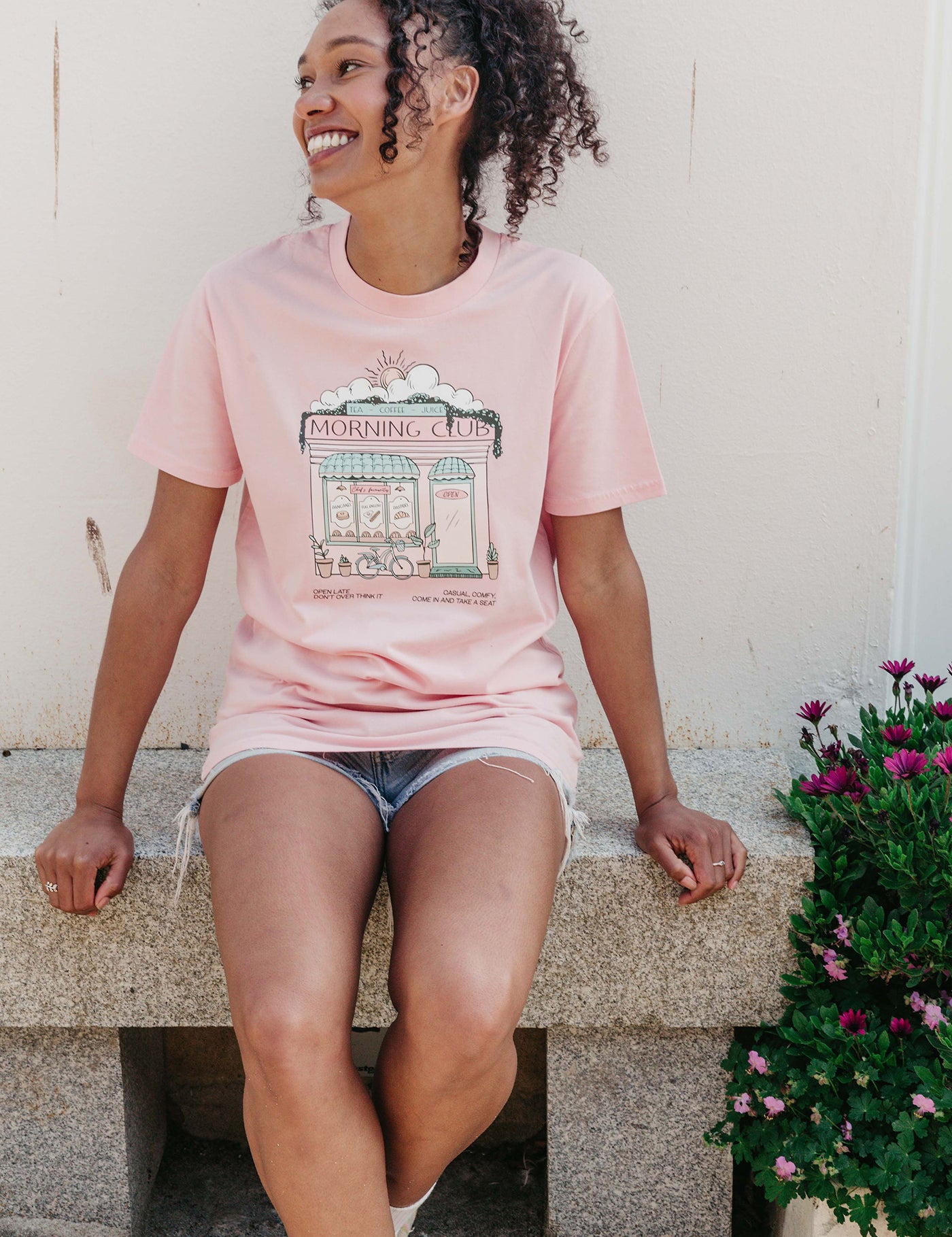 A lady wearing a pink printed graphic t-shirt, Morning Club Cafe, set in a town.