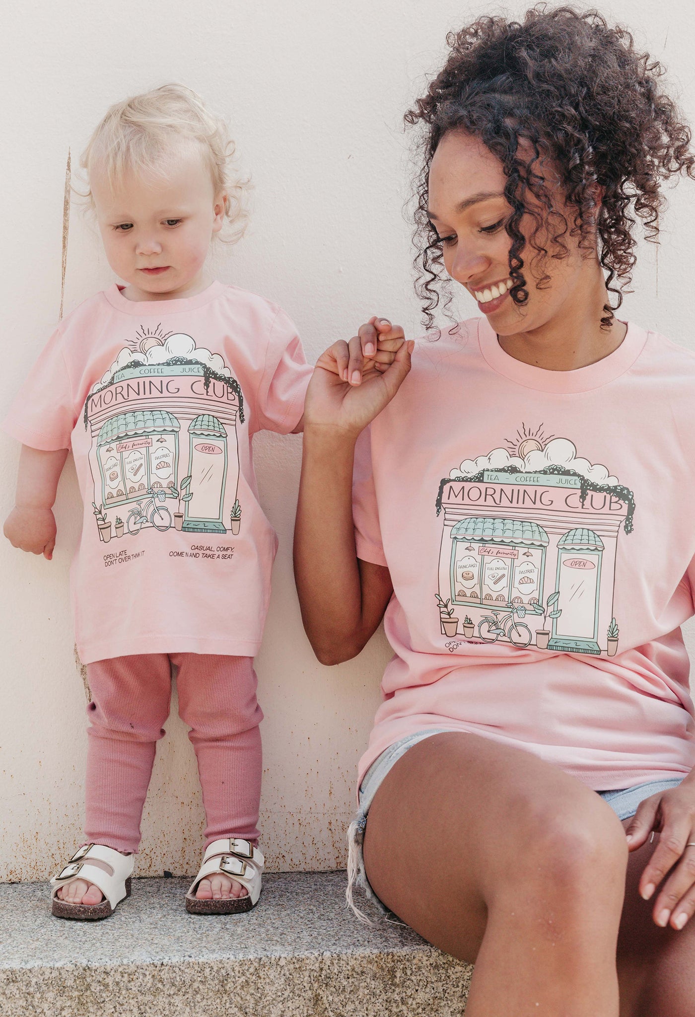 A lady and baby wearing a pink printed graphic t-shirt, Morning Club Cafe, set in a town.