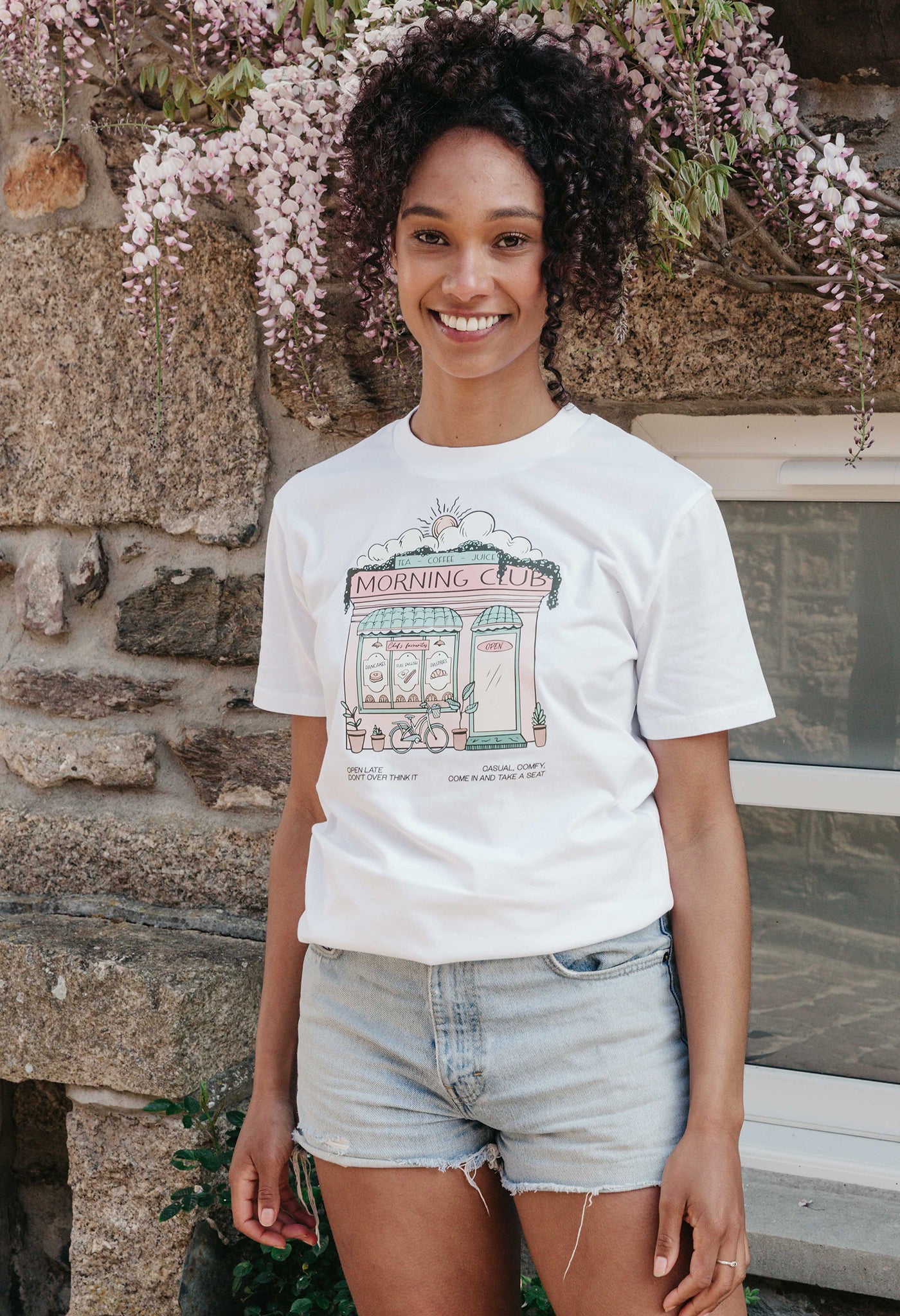 A lady wearing a white printed graphic t-shirt, Morning Club Cafe, set in a town.