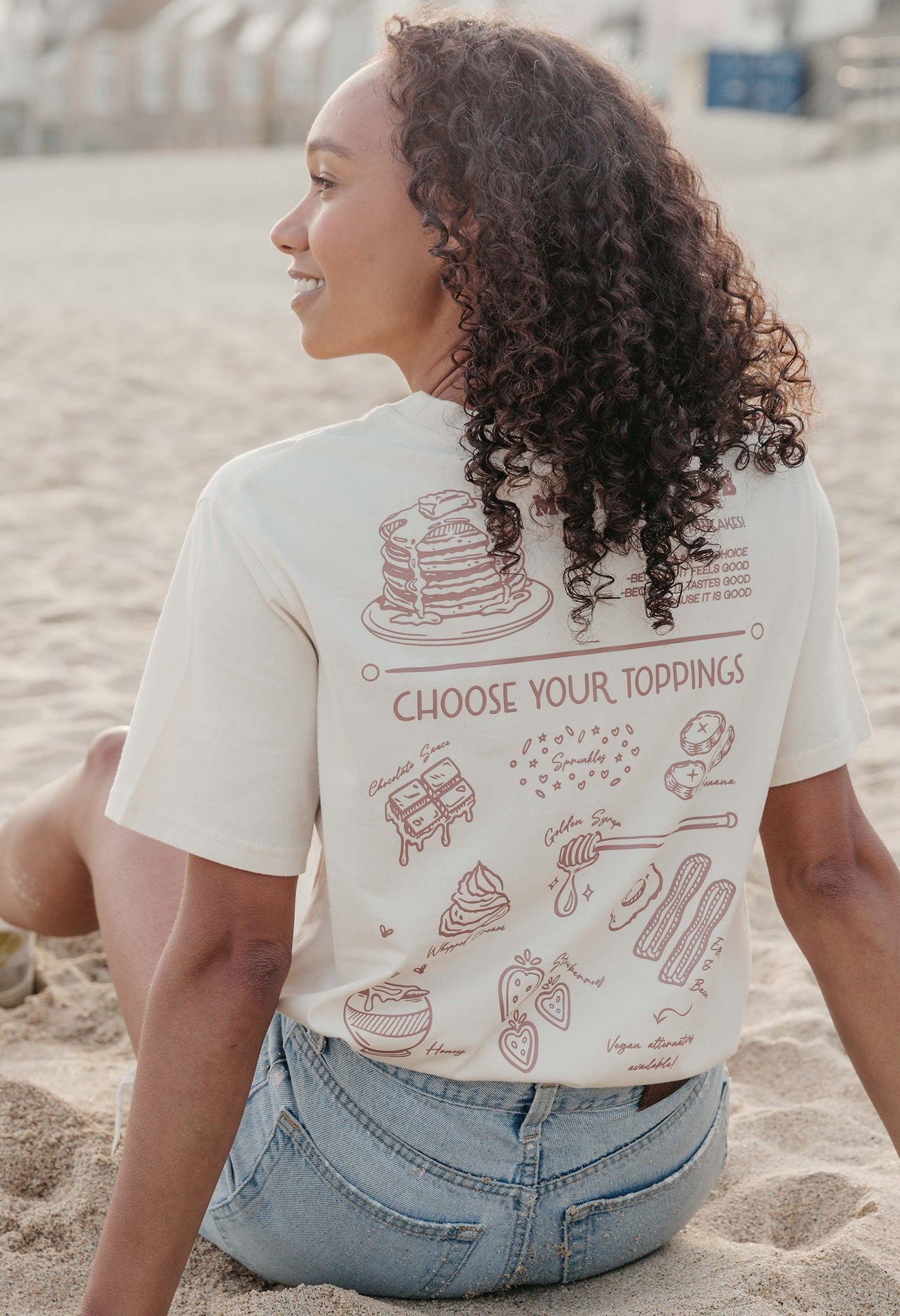 A lady wearing a natural printed graphic t-shirt, with a illustration of a pancakes and toppings.