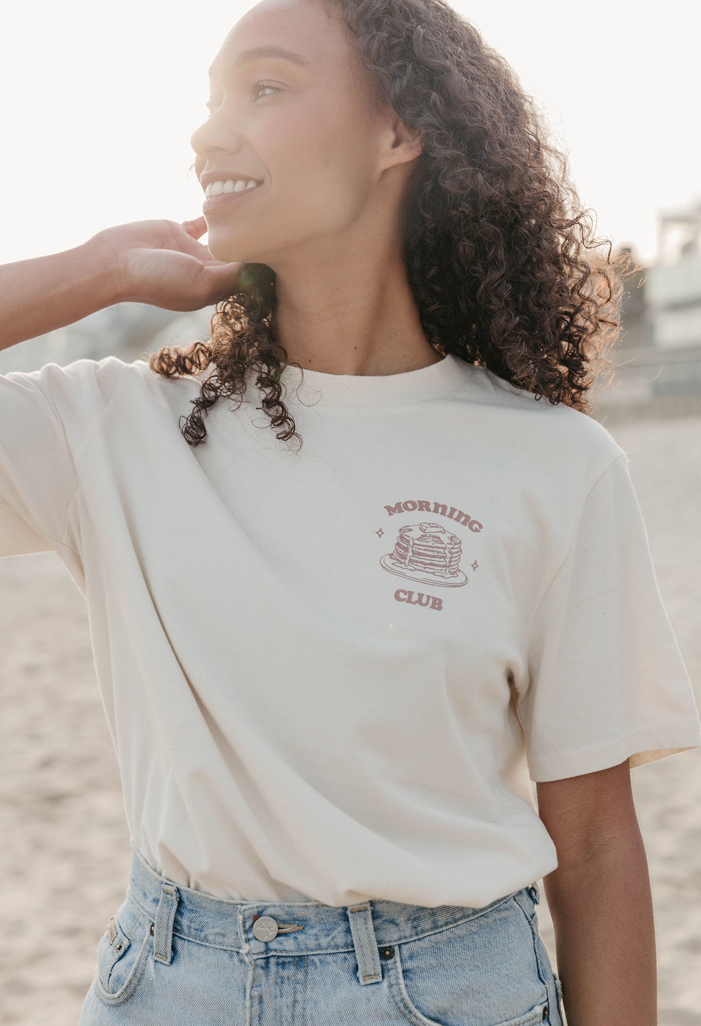 A lady wearing a natural printed graphic t-shirt, with a illustration of a pancakes and toppings.