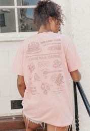 A lady wearing a pink printed graphic t-shirt, with a illustration of a pancakes and toppings.