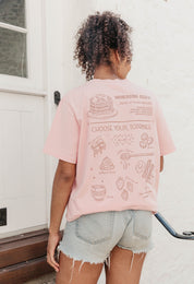 A lady wearing a pink printed graphic t-shirt, with a illustration of a pancakes and toppings.