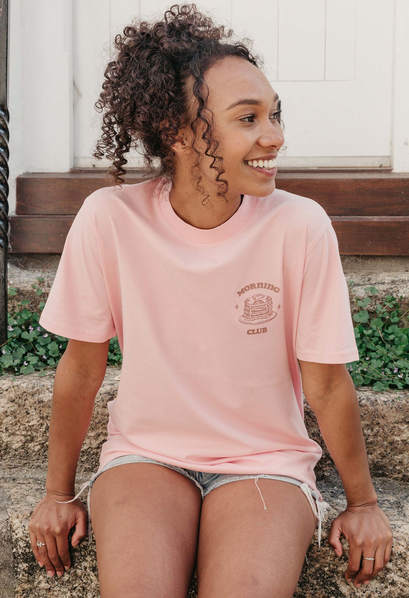 A lady wearing a pink printed graphic t-shirt, with a illustration of a pancakes and toppings.