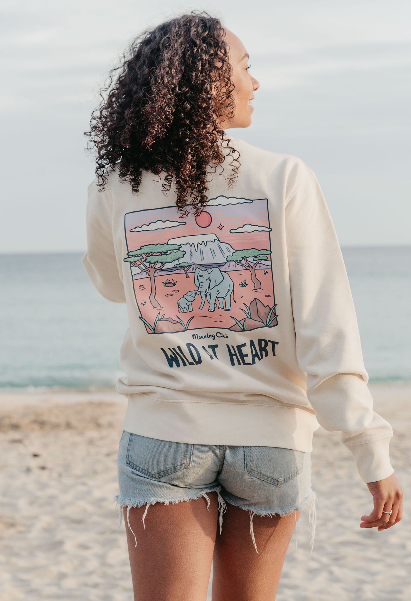 Lady wearing a printed sweatshirt, that says wild at heart, and its set on the beach.