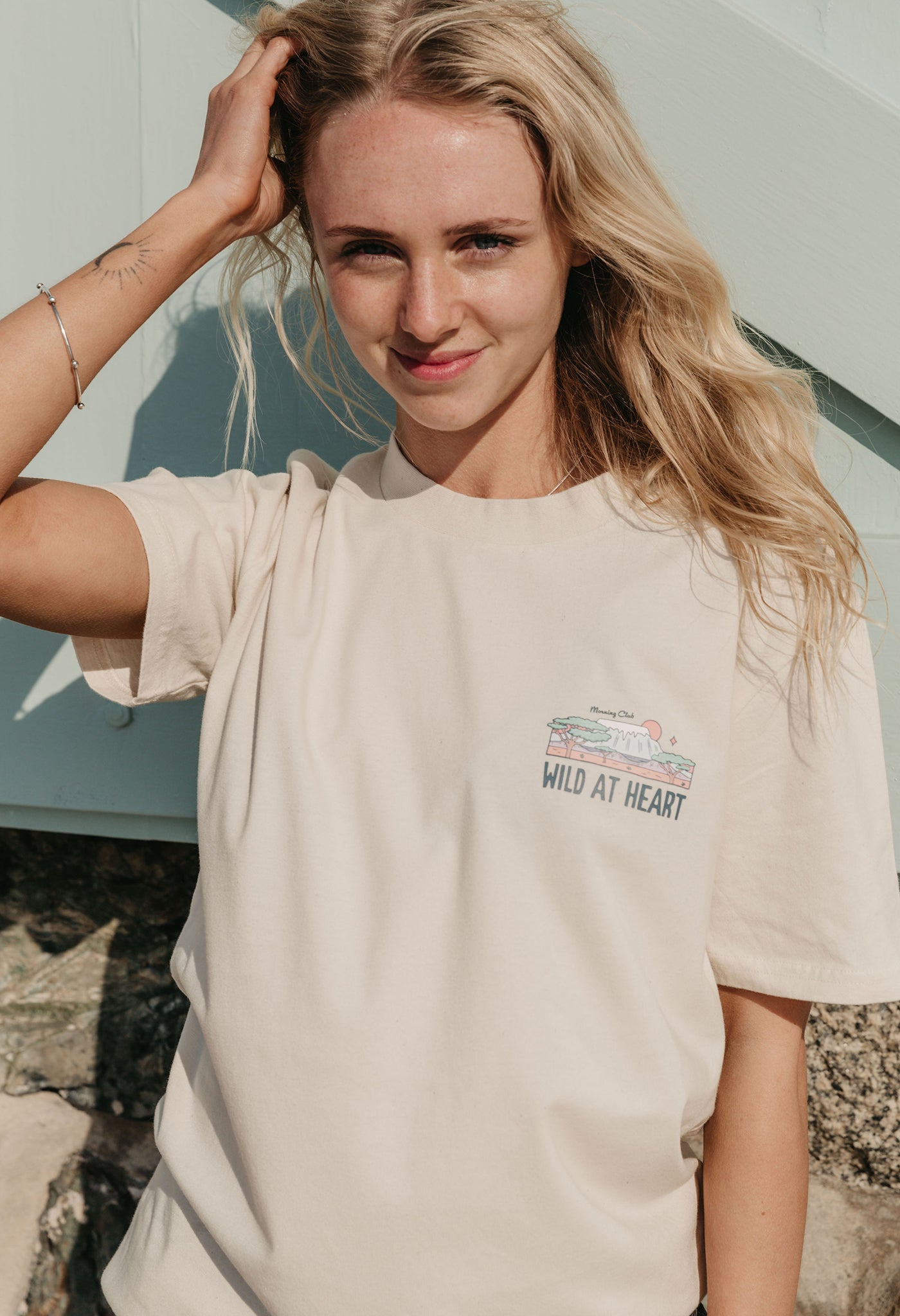 A lady wearing a natural printed graphic t-shirt, that says wild at heart, set by the beach.