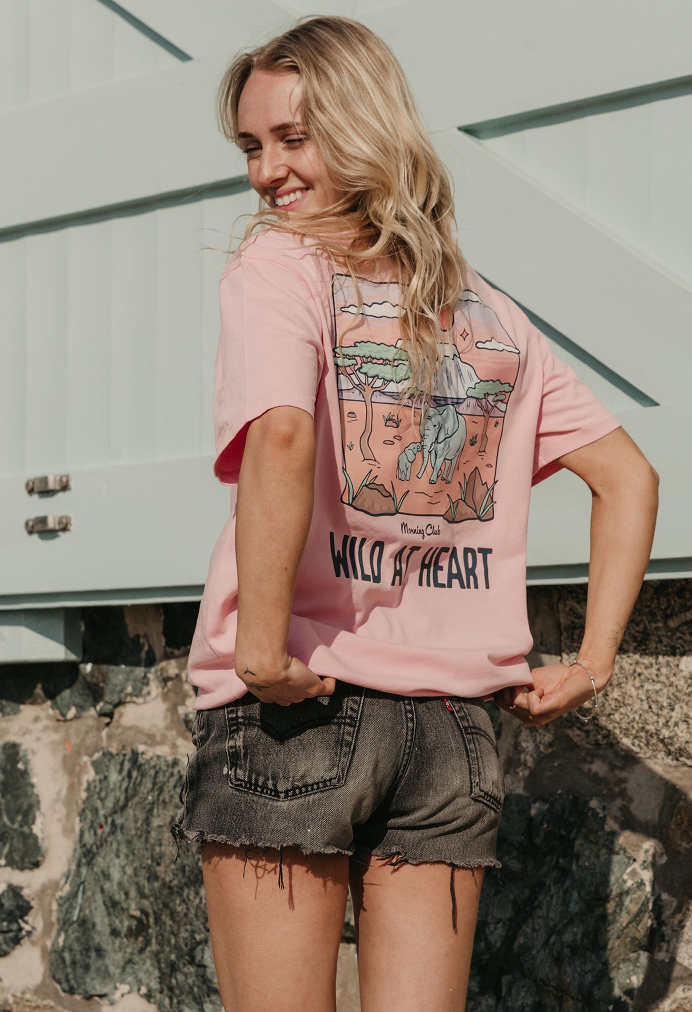 A lady wearing a pink printed graphic t-shirt, that says wild at heart, set by the beach.