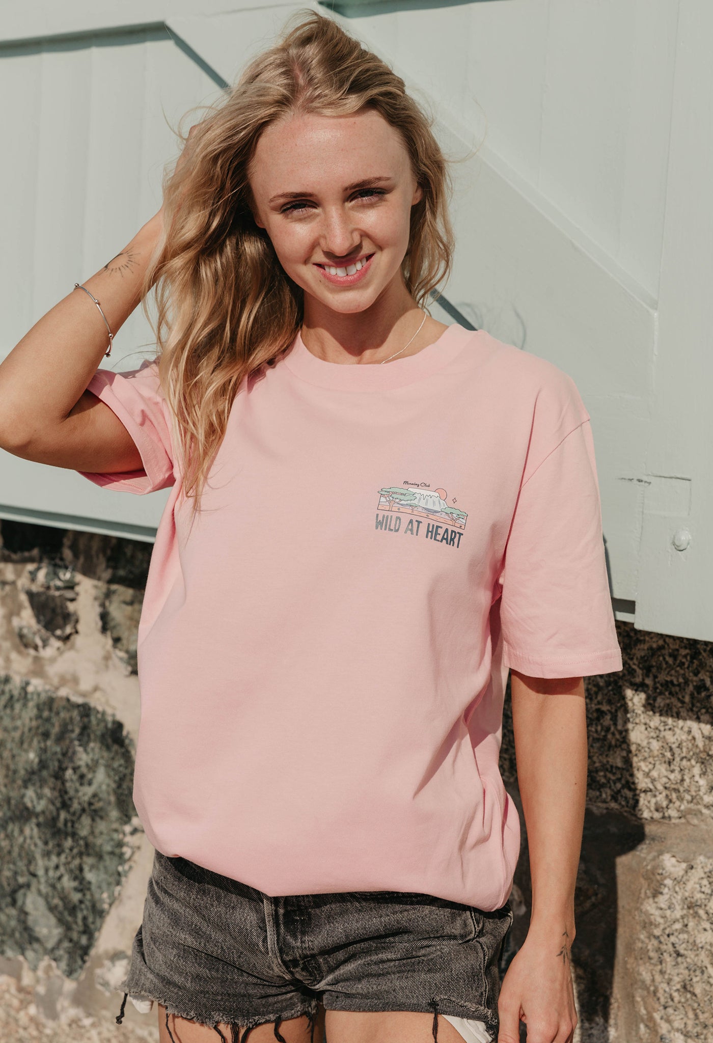 A lady wearing a pink printed graphic t-shirt, that says wild at heart, set by the beach.