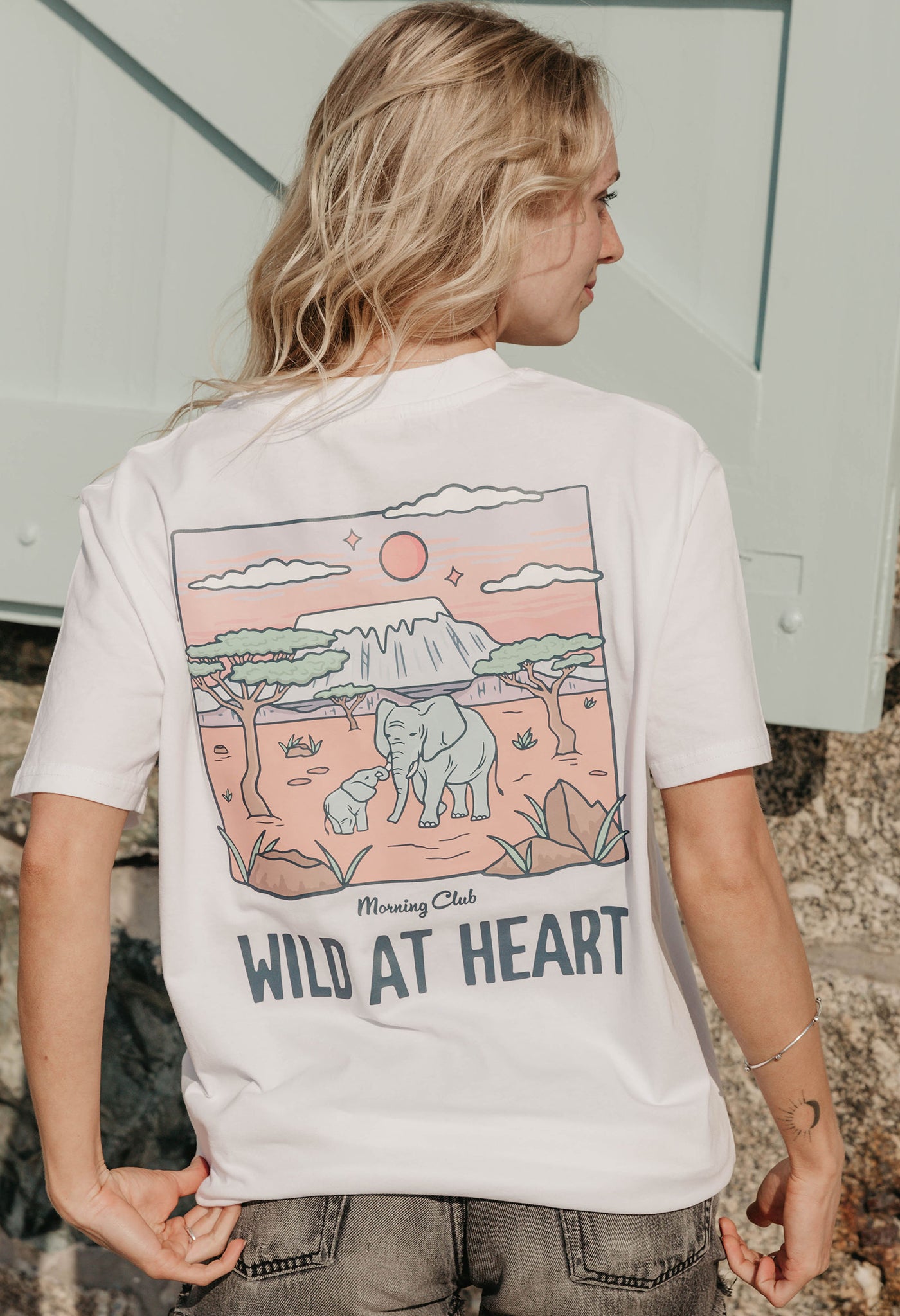 A lady wearing a white printed graphic t-shirt, that says wild at heart, set by the beach.