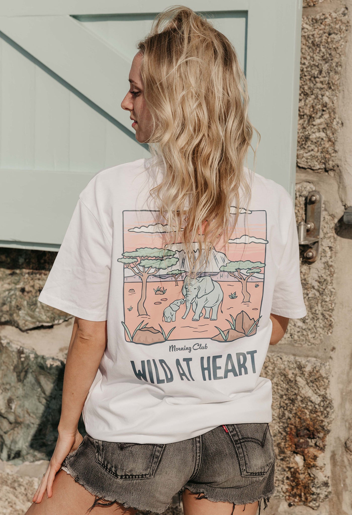 A lady wearing a white printed graphic t-shirt, that says wild at heart, set by the beach.