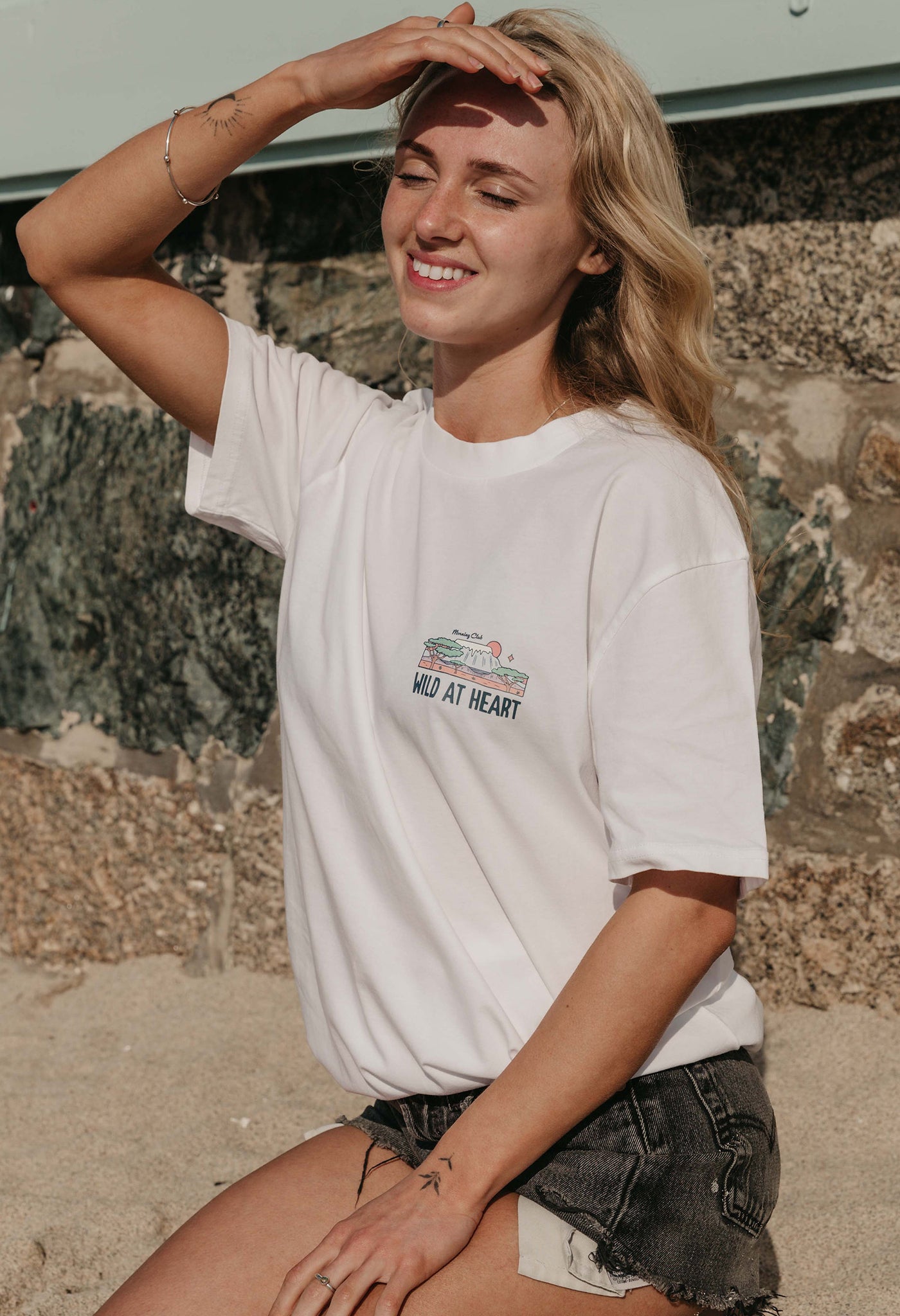 A lady wearing a white printed graphic t-shirt, that says wild at heart, set by the beach.