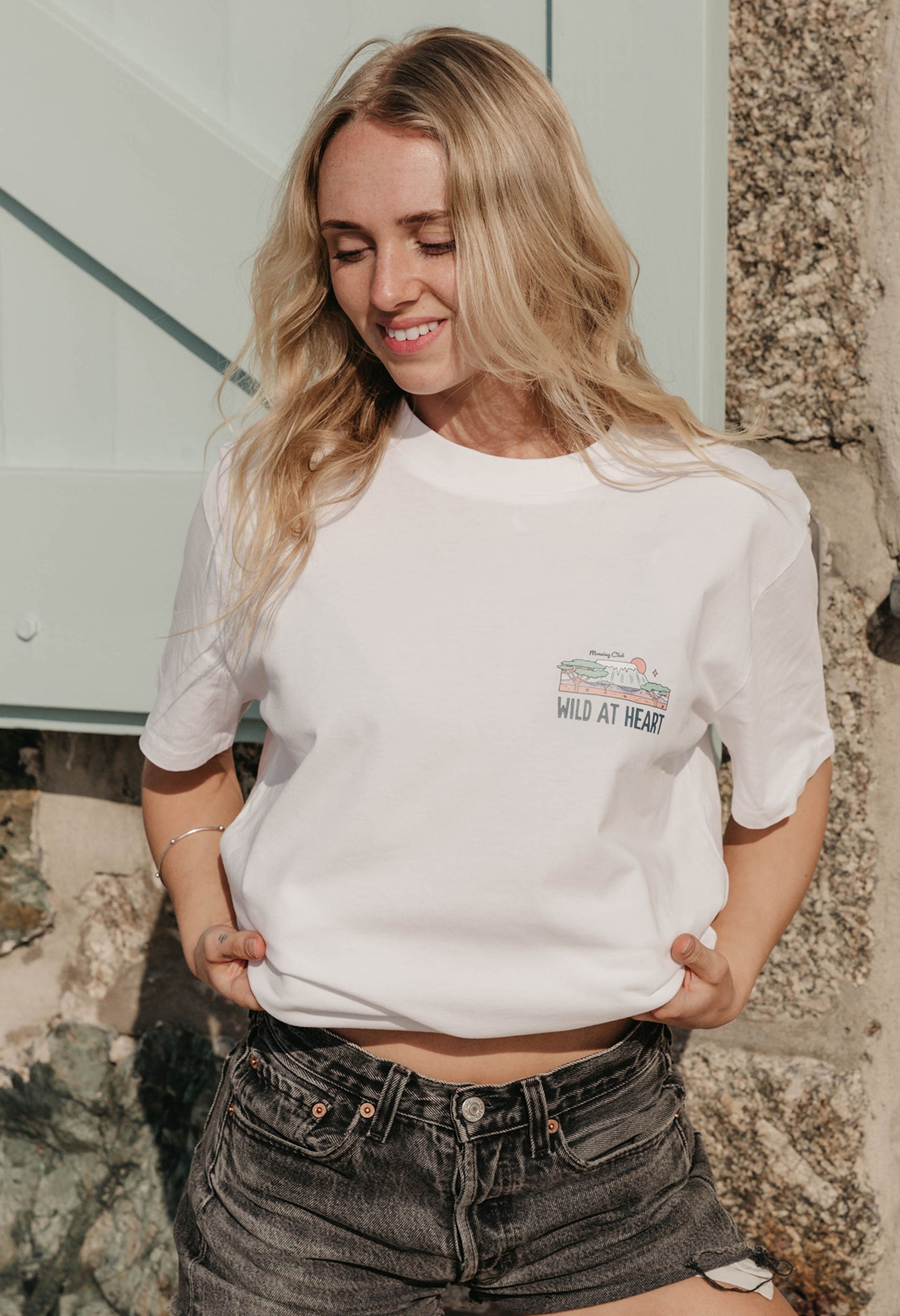 A lady wearing a white printed graphic t-shirt, that says wild at heart, set by the beach.